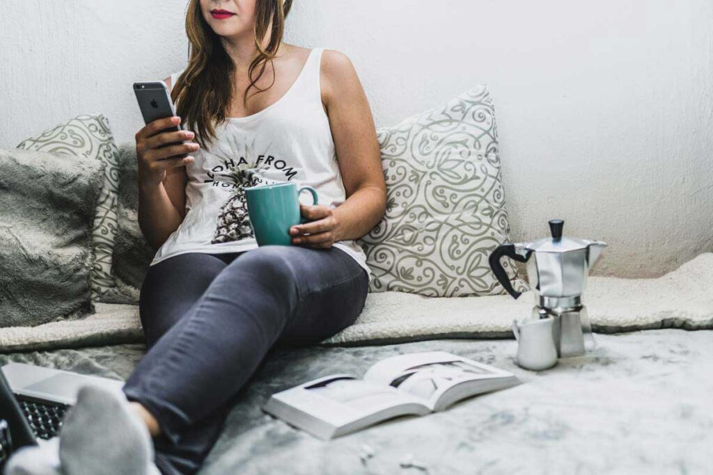 Woman lies comfortably on bed with a coffee and uses her phone