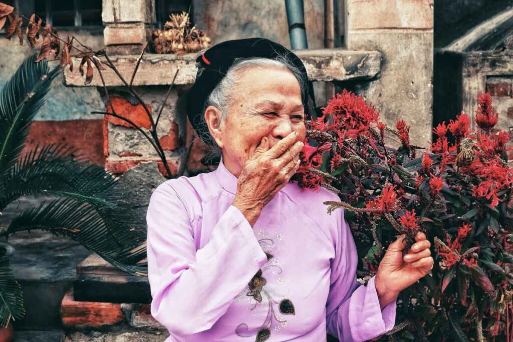 An elderly woman laughs in front of red flowers 