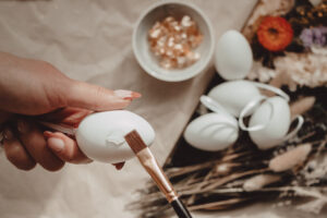 A pain brush draws on glue in a pattern. In the background the gold leaf and 4 plain eggs are visable.