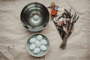 A flat lay view of a bowl of water next to a bowl of Easter eggs and a bunch of dried flowers