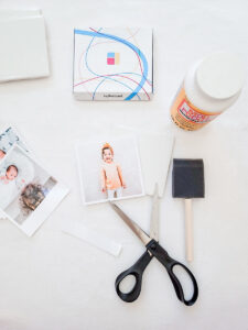Scissors next to a cut out photo surrounded by the materials for the DIY coaster: a ceramic tile, more MyPostcard photos, Mod Podge and a brush.