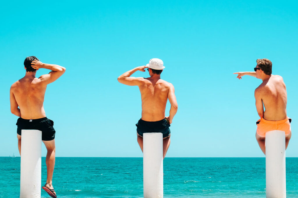 3 Männer sitzen auf Stöcke vor einem blauen Ozean auf dem Strand. aud
