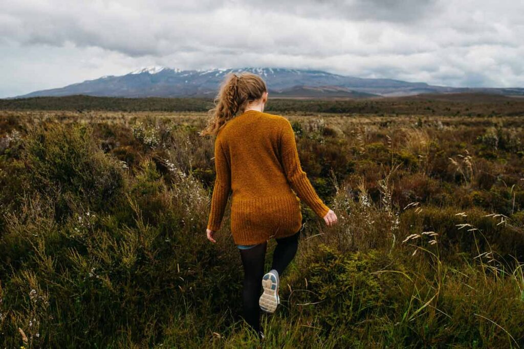 Eine Frau lauft weg durch einem Feld. Im Hintergrund sieht man Berge.