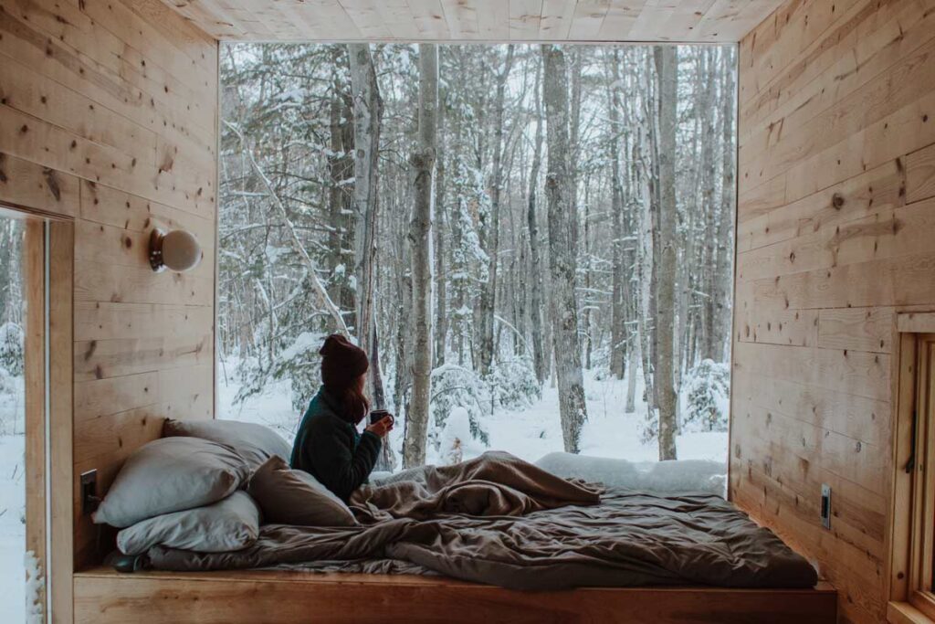 A woman sits on the bed in one of our places to go glamping and looks at the snowy woodland outside.