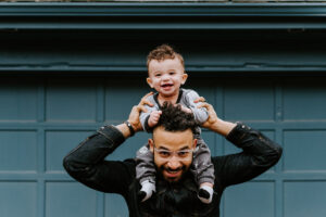 A man with his toddler sitting on his shoulders in front of a teal blue garage