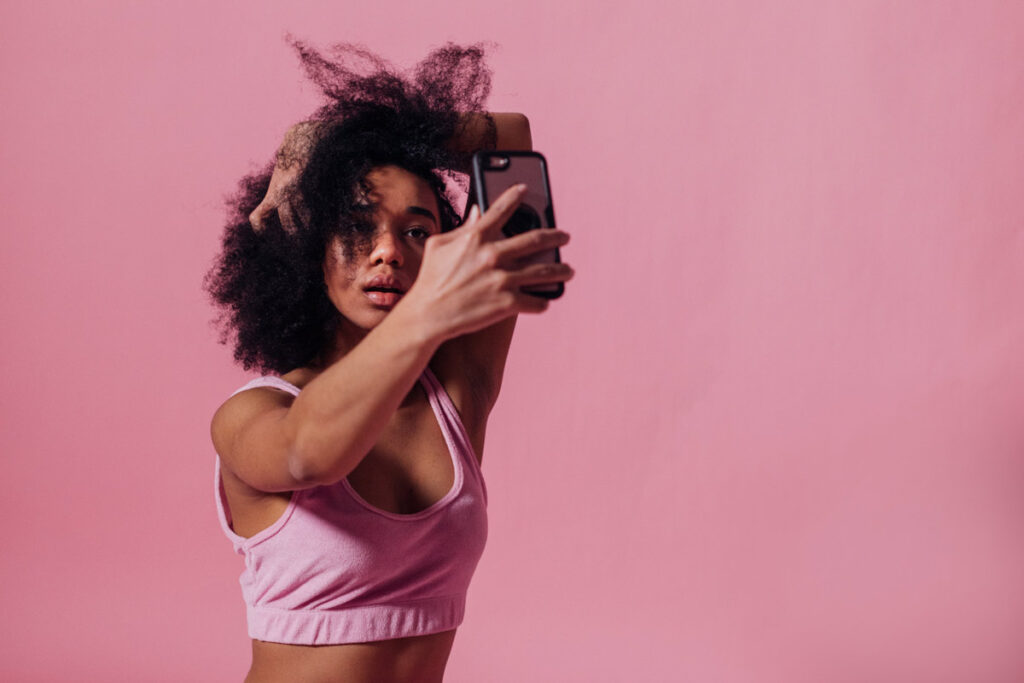 A woman standing in a pink room with a punk crop top on takes a sexy selfie, posing with her hand in her hair and mouth parted.