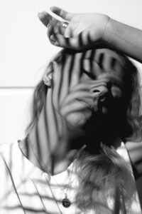 A woman in black and white with bar shadows across the face, poses with her hand over her head and eyes closed.