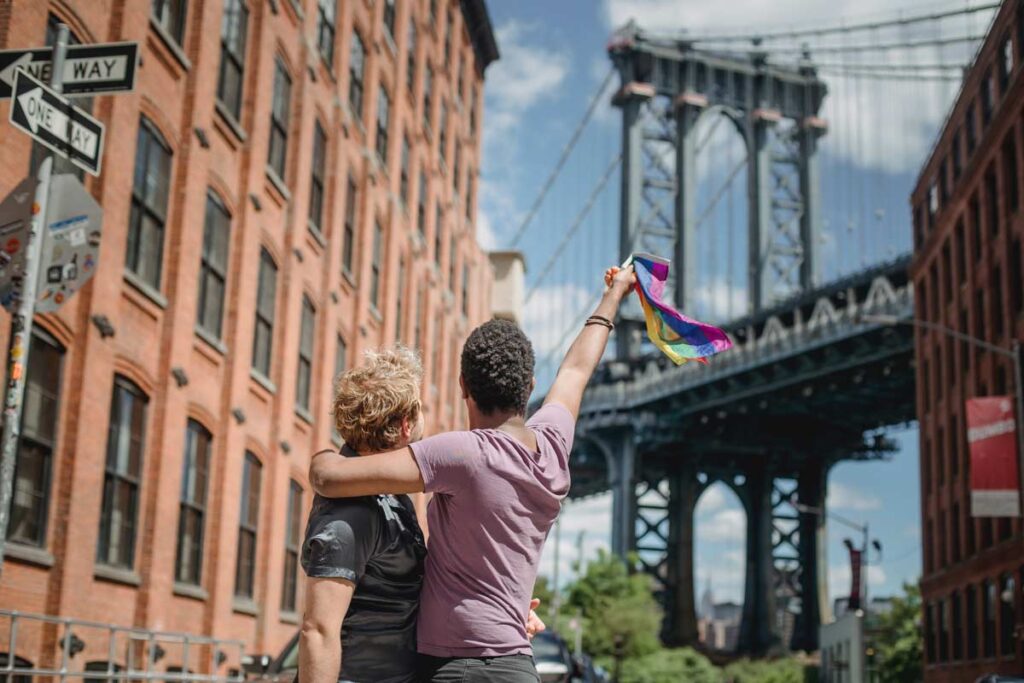 A couple from the LGBTQ+ community hug each other in a city scape backs to camera waving a rainbow flag.