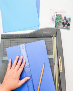 A piece of blue paper is being cut on top of a cutter while a hand rests on top next to a ruler.