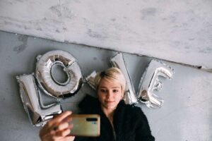 Taking a selfie from above, a woman lies on the ground on top of balloons shaped to spell LOVE