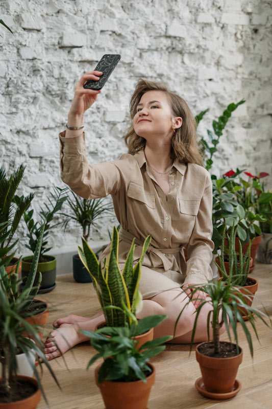 A woman takes a selfie portrait from above while sitting surrounded by plants on the floor.