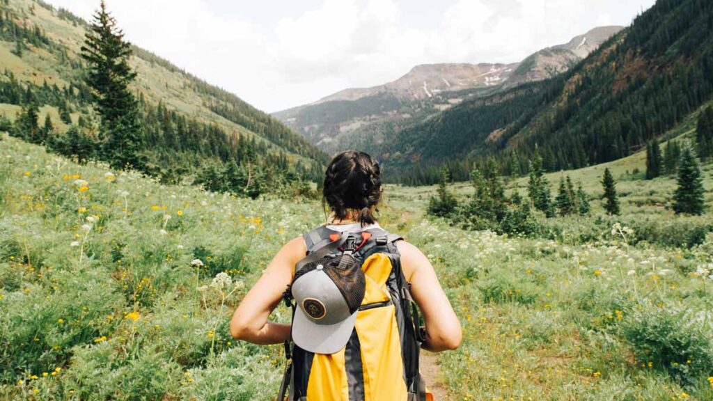 A woman with a yellow hiking backpack is on a trail between a valley