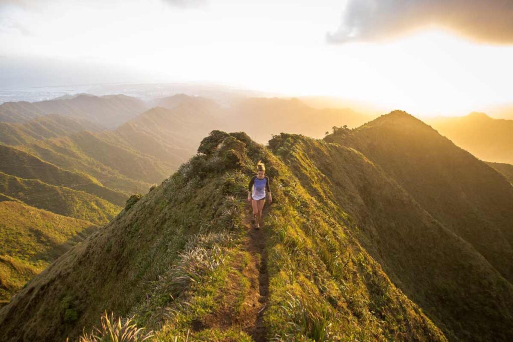 At the top of a mountain, a woman ahead hiking in the setting sun