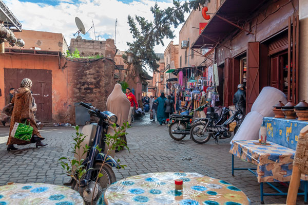 Eine Gasse in Marrakesch. Zu sehen sind Menschen, die passieren, und vereinzelt Tische, die auf der Straße stehen.