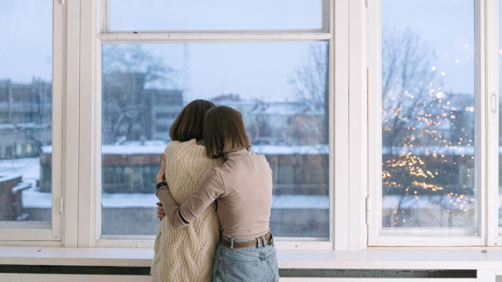 Two women hug in front of a window