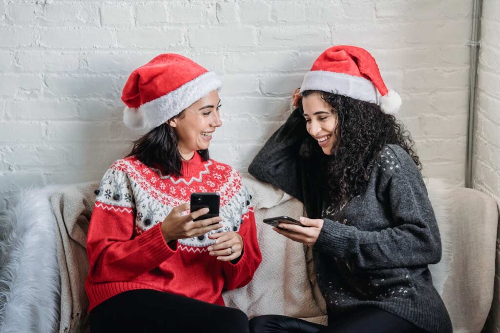Two women on the couch with christmas hats and sweaters on Instagram and laughing