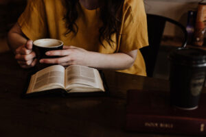 Zu sehen sind die Hände einer Frau mit Kaffee in der Hand. Vor ihr liegt ein Buch. Sie liest.