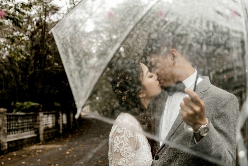 A couple kiss under a clear umbrella for their creative pre wedding photoshoot