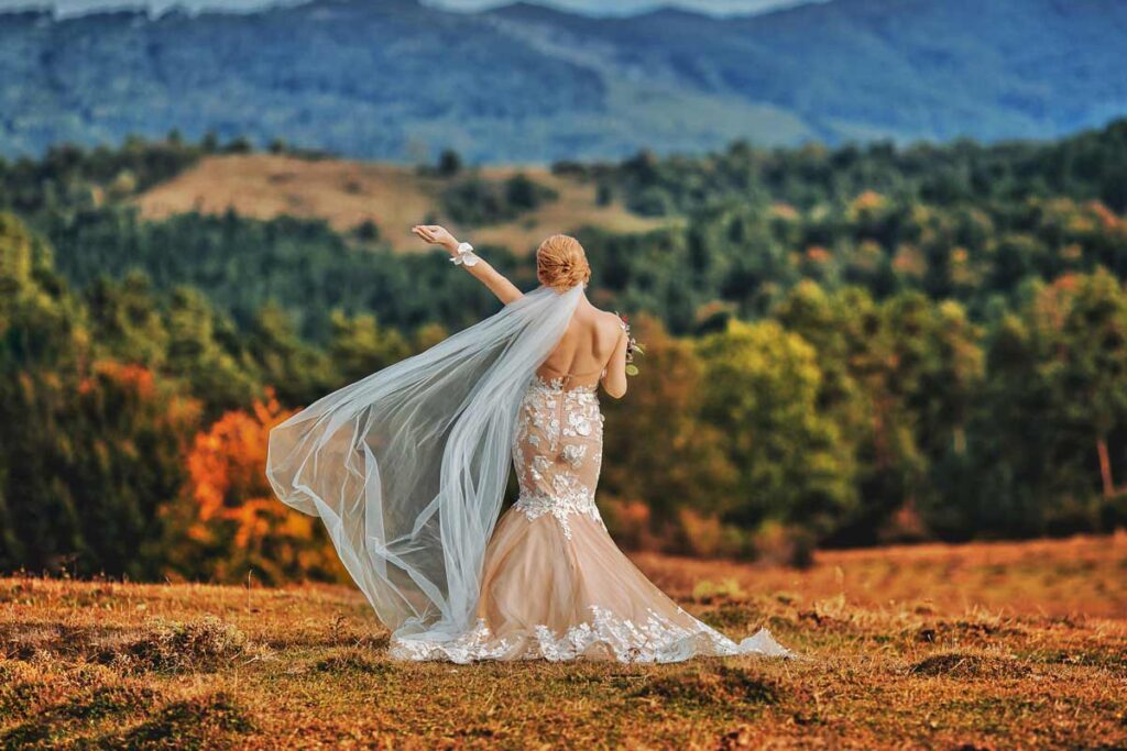 A bride stands facing the wooded landcape in her wedding dress, posing with arm up