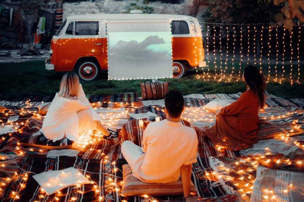 A group of 3 sit on a rug in front of a projection in front of a car