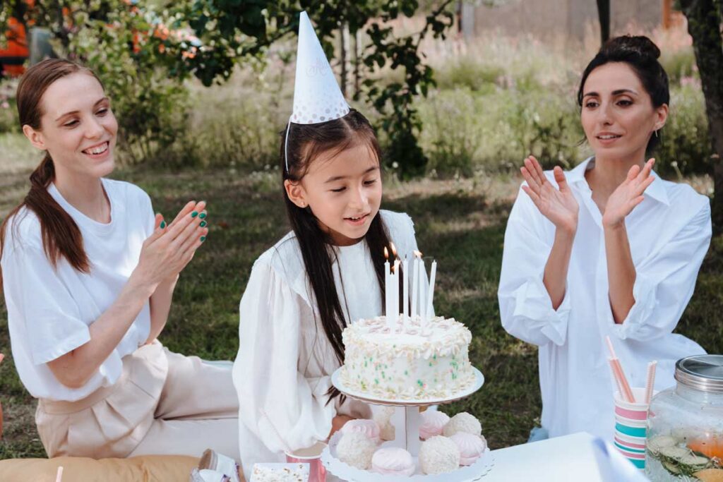 A young girl blows with a party hat blows out candles on cake next to two young adults