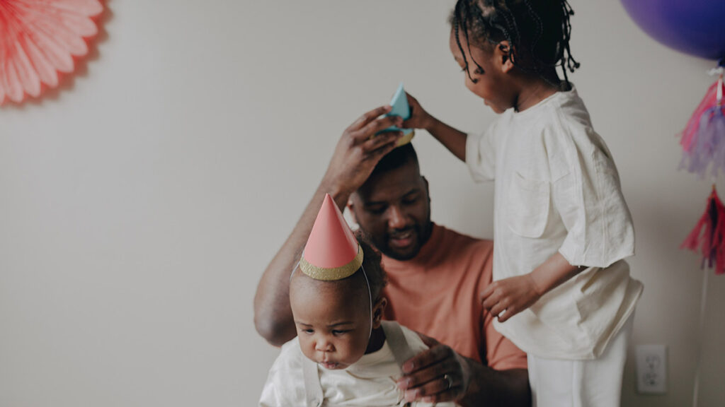 A father's son puts a party hat on his head as his toddler sits on his lap.