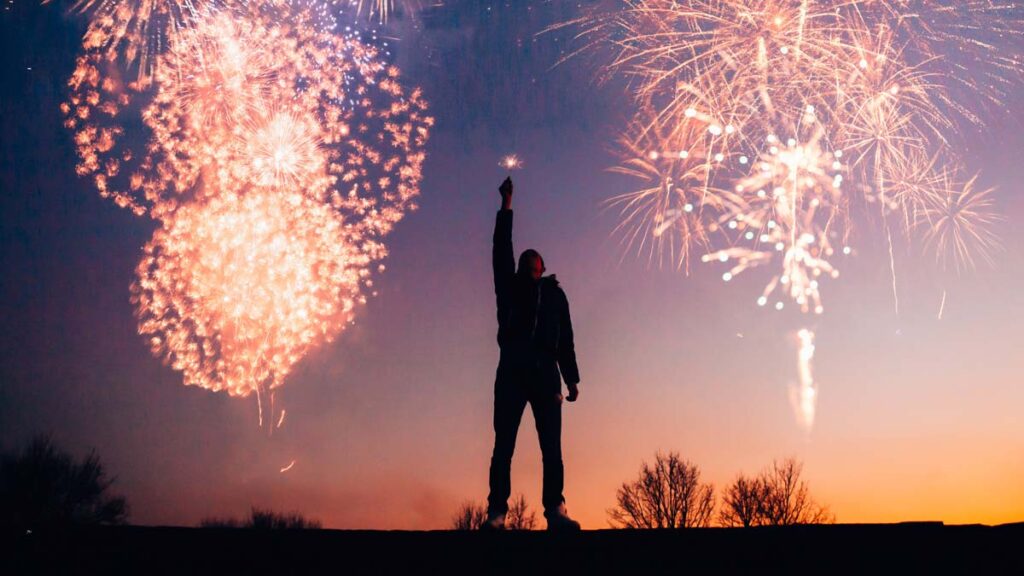 ein Mann hält eine Wunderkerze in die Luft, während über ihm Feuerwerke zu sehen sind