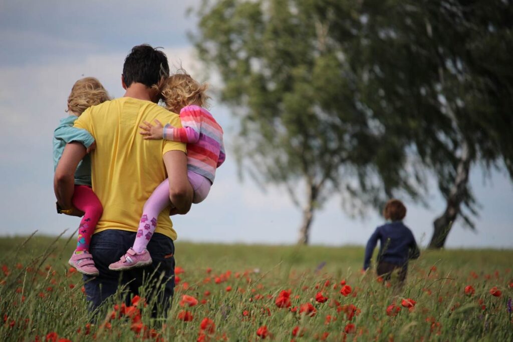 A father holding two kids in a field of flowers while a third runs ahead.